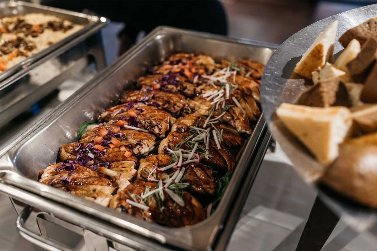 millcreek-img-3 A metal buffet tray filled with cooked chicken breast topped with vegetables and herbs, with a side dish visible on the right.