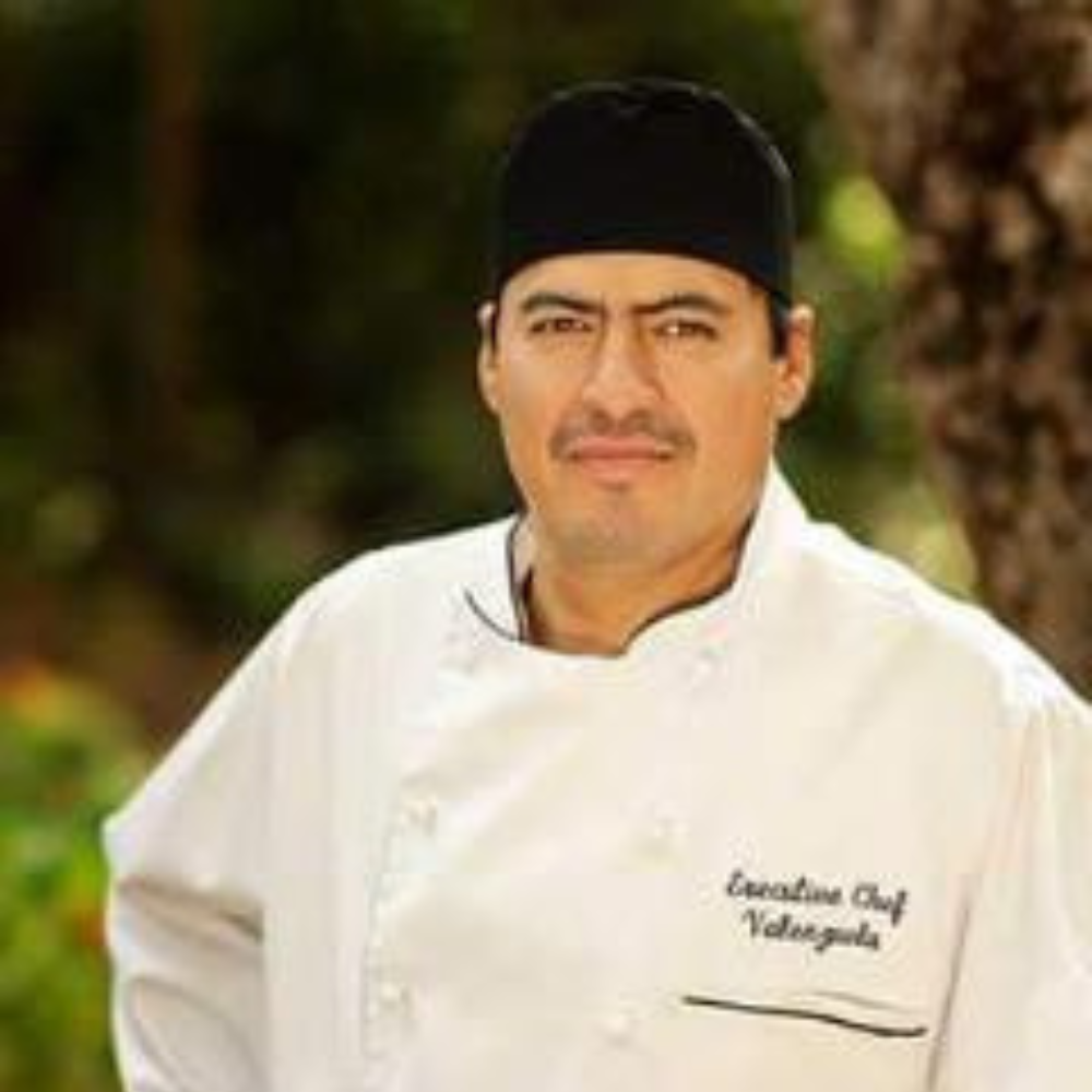 Untitled design (1) copy 2 A man wearing a chef's uniform and black chef's hat stands outdoors with trees in the background. He has a serious expression and his hands are on his hips. The uniform reads "Executive Chef Velazques".